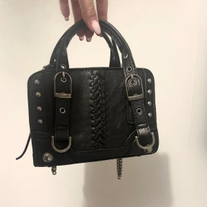 Svart handväska med nitar  - Snygg svart handväska i skinn med coola kedjedetaljer och nitar. Väskan har en flätad design på framsidan och justerbara remmar med spännen. Perfekt för en rockig look eller en utekväll. Den har också en dragkedja på sidan för extra säkerhet. Även ett mindre fack på insidan.