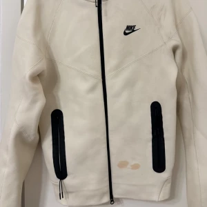 Vit kofta från Nike - Säljer en vit Nike-kofta med svarta detaljer och dragkedja. Den har en huva och två fickor framtill. Några små fläckar finns på framsidan, som ej går bort i vanlig tvätt, skulle dock säga att man kan testa använda vanish eller klorin, vilket jag inte testat göra då jag köpte en ny.❤️Använt fåtal gånger och köpt från Zalando