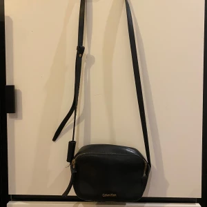 Svart axelremsväska från Calvin Klein - Snygg svart axelväska från Calvin Klein i skinn. Väskan har en justerbar rem och en praktisk dragkedja upptill. Perfekt för både vardag och fest! 🖤