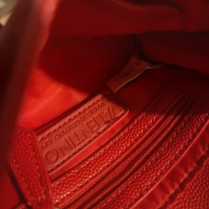 Röd axelväska från Valentino - Snygg röd axelväska från Valentino i skinn med en elegant kedjerem. Väskan har en stilren design med ett V-emblem på framsidan och stängs med ett trycklås. Perfekt för både vardag och fest!