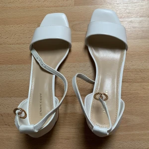Vita sandaler med klack från Jenny Fairy - Snygga vita sandaler från Jenny Fairy i nytt skick. De har en elegant klack och en justerbar rem med gulddetaljer. Perfekta för sommarens fester eller en dag på stan. Stilrena och bekväma att bära hela dagen.