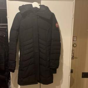 Säljer en svart lång dunjacka från Canada Goose. Perfekt för kalla vinterdagar med sin varma och isolerande design. Jackan har en huva och långa ärmar, samt en dragkedja framtill. Den är i bra skick och kvitto finns