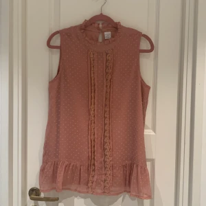 Rosa ärmlös blus från Lindex - Säljer en söt rosa ärmlös blus från Lindex i storlek L. Blusen har fina spetsdetaljer framtill och en volangkant längst ner. Den är perfekt för vår och sommar och har en knapp i nacken för enkel påklädning. Superfin till både jeans och kjol!