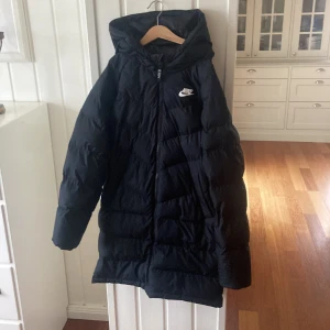 Svart dunjacka från Nike - Säljer en svart dunjacka från Nike i storlek 147-158cm Jackan har en lång och varm design med huva och dragkedja framtill. Perfekt för kalla vinterdagar. Den är i mycket bra skick och redo för nya äventyr! 🖤