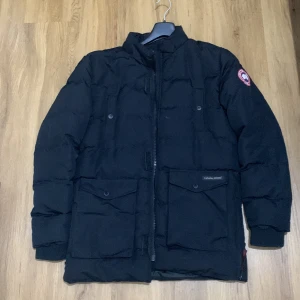 Svart dunjacka från Canada Goose - Säljer en svart dunjacka från Canada Goose i mycket bra skick. Jackan har en klassisk design med lång ärm och dragkedja framtill. Perfekt för kalla vinterdagar med sin varma och skyddande stil. Märkeslogga på ärmen ger en extra touch av stil. Passar perfekt för både vardag och utomhusaktiviteter.