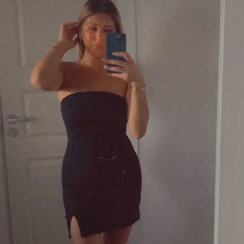 Snygg svart axelbandslös klänning i tight passform. Perfekt för festliga tillfällen med en stilren design och en liten slits framtill. Klänningen är kort och ger en elegant look.. Mekot.