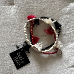 Armband från Bud to Rose - Säljer ett set med tre armband från Bud to Rose. De är i färgerna svart, vitt och rött med små pärlor och tofsar som detaljer. Perfekt för att ge en bohemisk touch till din outfit. Armbanden är elastiska och passar de flesta handleder. Helt nytt!