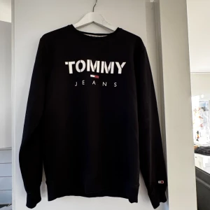 Svart sweatshirt från Tommy Jeans - Säljer en svart sweatshirt från Tommy Jeans i mycket bra skick. Den har en klassisk design med logotypen tryckt i vitt på bröstet. Perfekt för en avslappnad stil och passar till alla tillfällen. Skön och stilren!