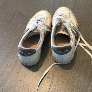 Vita sneakers från Veja - Säljer ett par vita sneakers från Veja i bra skick. De har en klassisk design med snörning och en grå detalj på hälen där märket syns tydligt. Perfekta för både vardag och festliga tillfällen. Storlek passar de flesta.