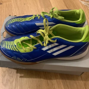 Blå och gröna fotbollsskor från Adidas - Säljer ett par coola Adidas fotbollsskor i blått och grönt. De har en snygg design med vita ränder och grön snörning. Perfekta för inomhusfotboll med en slitstark sula. Skorna är i bra skick och redo för nya äventyr på planen!