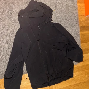 Svart vindjacka från Nike - Säljer en svart vindjacka från Nike i mycket bra skick. Jackan har en dragkedja framtill och en praktisk huva. Perfekt för höst och vår när det blåser lite extra. Den är långärmad och har en stilren design som passar till det mesta. Perfekt för både träning och vardag! Köpt för 1299kr. Storlek L men passar M väldigt bra 