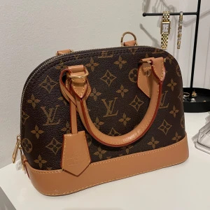 Brun handväska - Super fin och söt handväska i mycket fint skick förutom att de finns en liten vit fläck som syns på bilden men inget man lägger märke till. Väskan är stilren och passar till alla årstider. Tillkommande band syns på sista bilden. Måtten är 18 x 25 cm.