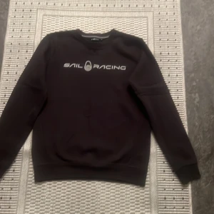 Svart tröja från Sail Racing - Säljer en svart tröja från Sail Racing i storlek S. Tröjan har en stilren design med Sail Racing-loggan tryckt på framsidan. Den är perfekt för både vardag och fritid. Materialet känns mjukt och bekvämt, och den har långa ärmar med ribbade muddar.