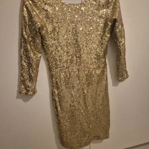Glittrig guldklänning - Snygg glittrig guldklänning perfekt för festliga tillfällen! Den har trekvartsärm och är täckt med paljetter som verkligen fångar ljuset. Klänningen är i ett tight passform och passar perfekt för nyår eller andra glamorösa fester. Gör ett statement med denna glittriga skönhet!