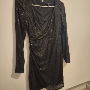Svart glittrig klänning - Snygg svart glittrig klänning perfekt för festliga tillfällen. Den har en omlott-design och långa ärmar som ger en elegant look. Klänningen är i mycket bra skick och passar perfekt för nyår eller andra festliga tillfällen. Den är gjord av ett bekvämt material som sitter bra på kroppen.