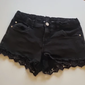 Svarta shorts som är lågmidjade med spetsdetaljer från Kappahl - Snygga svarta shorts från Kappahl i storlek 158. De har en cool spetsdetalj längst ner och är perfekta för sommaren. Shortsen har fickor fram och bak samt en knapp och dragkedja framtill. Perfekta för en avslappnad men stilren look!