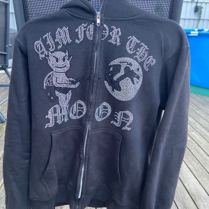Svart hoodie med tryck - Säljer en svart hoodie med coolt tryck 'AIM FOR THE MOON' och en figur på framsidan. Den har dragkedja och är i bra skick. Perfekt för höst och vinter, och passar till vardags eller chill dagar. Storleken är M och denna snygga tröja kommer tyvärr inte till användning längre