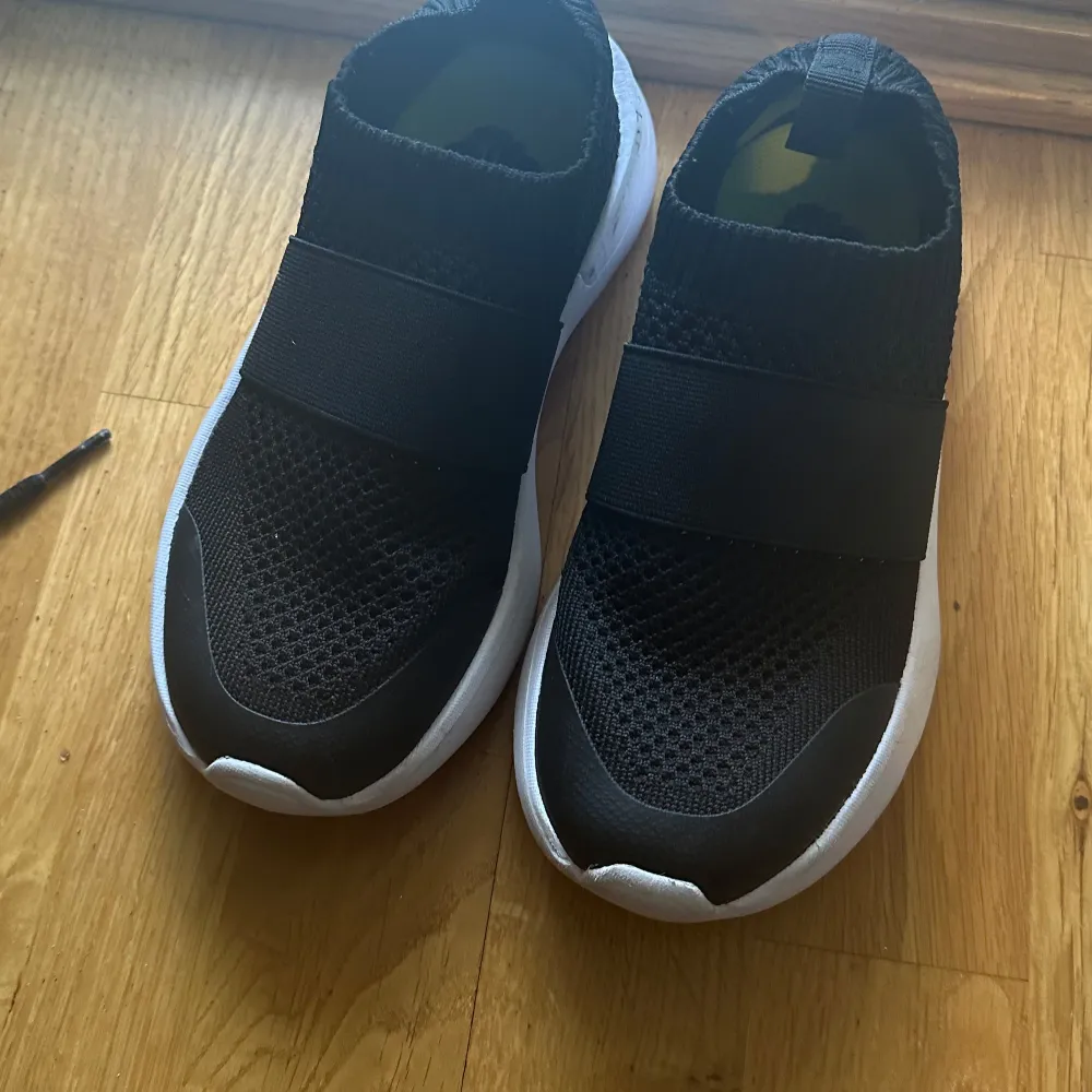 Säljer ett par svarta sneakers i mycket bra skick. De har en elastisk rem över foten för en tight passform och en vit sula som ger en snygg kontrast. Perfekta för både träning och vardagsbruk. Storlek är inte specificerad, men de ser ut att vara i normal passform.. Skor.