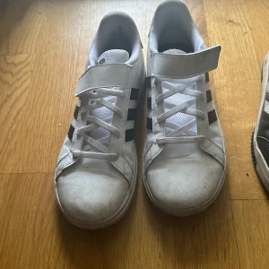 Vita Adidas sneakers med svarta ränder - Säljer ett par klassiska vita Adidas sneakers med svarta ränder. De har snörning och kardborreband för extra säkerhet. Skorna är i acceptabelt skick med lite smuts på tån. Perfekta för vardagsbruk och passar till det mesta! man behöver bara tvätta och bli fint och ny 