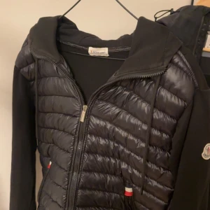 Moncler cardigan - Säljer en snygg Moncler cardigan. Den har en quiltad framsida med dragkedja och en huva för extra stil och värme. Perfekt för höst och vår! Jackan har Moncler-loggan på ärmen och är i mycket bra skick. Köpt för 2500kr här på plick!