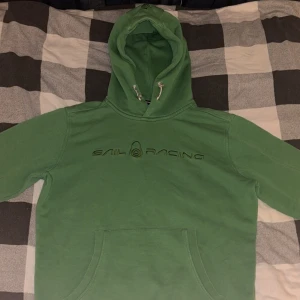 Grön hoodie från Sail Racing - Säljer en snygg grön hoodie från Sail Racing i storlek S. Den har en klassisk design med känguruficka och justerbar huva med snören. Perfekt för både vardag och chill. Den är i bra skick och redo för nya äventyr! 💚