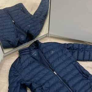 Mörkblå pufferjacka från GANT - Säljer en snygg mörkblå pufferjacka från GANT i storlek medium. Jackan har en dragkedja framtill och är perfekt för kyliga dagar. Den är i mycket bra skick och passar både till vardags och lite finare tillfällen. Perfekt för höst och vinter!