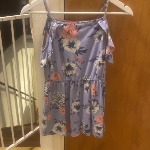 Blommig sommarklänning - Söt blommig klänning i ljuslila med rosa och vita blommor. Den har smala axelband och en avslappnad passform, perfekt för varma sommardagar. Klänningen är kort och har en luftig design som gör den idealisk för både strand och stad.