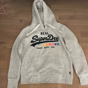 Grå hoodie från Superdry - Säljer en snygg grå hoodie från Superdry i bra skick. Den är lite liten i storleken så jag skulle säja att den är mer i storlek 38