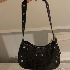 Svart axelväska med krokodilmönster - Snygg svart axelväska i syntet med krokodilmönster och silverdetaljer. Väskan har en dragkedja upptill och en extra ficka med dragkedja på baksidan. 