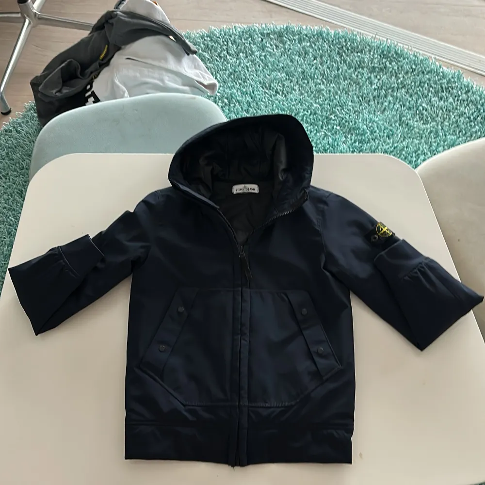 Säljer en snygg blå jacka från Stone Island Junior. Den har en dragkedja framtill och långa ärmar. Perfekt för höst och vår. Jackan är i mycket bra skick och har en cool logga på ärmen. Passar perfekt för dig som vill ha en stilren look!. Takit.