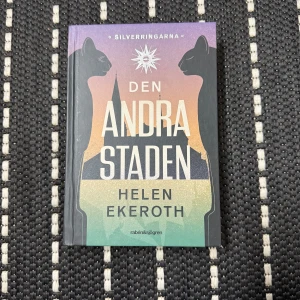 Den andra staden - Säljer boken 'Den andra staden' av Helen Ekeroth. Det är första delen i serien Silverringarna. Boken har en mystisk och färgglad omslagsdesign med silhuetter av katter och en stad. Perfekt för dig som gillar fantasy och äventyr! Helt oanvänd, köpt för 199kr, säljer för 100kr 📚✨ 