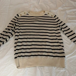 Randig tröja med knappar - Säljer en snygg randig tröja  i beige och svart med coola knappar på axlarna.Från HM I storlek M Den är långärmad och har ribbade muddar vid ärmslut och nederkant. Perfekt för en stilren look i vår och höst!