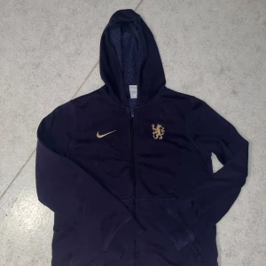 Mörkblå hoodie från Nike - Säljer en snygg mörkblå hoodie från Nike med dragkedja och huva. Den har ett guldigt lejonemblem på bröstet och Nike-logga. Perfekt för höst och vinter, håller dig varm och stilren. Passar både till vardags och träning.