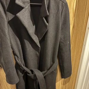 Svart kappa med bälte - Säljer en elegant svart kappa i mycket bra skick. Den har ett bälte i midjan för en snygg passform och långa ärmar. Perfekt för höst och vår när du vill hålla dig varm och stilren. Passar både till vardags och festliga tillfällen.