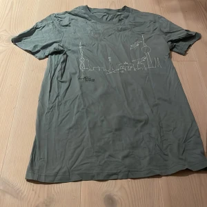 Grön t-shirt med stadssilhuett - Säljer en grön t-shirt i bomull med en cool stadssilhuett tryckt på framsidan. Perfekt för vardagsbruk eller en avslappnad dag ute. T-shirten är i bra skick och har korta ärmar. Passar perfekt för sommaren eller våren!