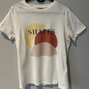 Vit t-shirt med grafiskt tryck - En vit t-shirt med ett grafiskt tryck i gult, rosa och beige. Perfekt för en avslappnad stil. T-shirten är kortärmad och i bomull, perfekt till vardagen. Storlek S, passar även M. Oanvänd. Skriv för fler bilder! 