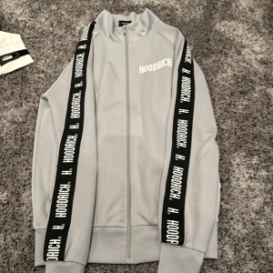 Grå kofta från Hoodrich - Säljer en snygg grå kofta från Hoodrich med dragkedja och svarta band med logga längs ärmarna. Byxorna till den finns skriv i dm. Köp båda för bättre pris. Perfekt för en casual look eller träning. Koftan har en hög krage och är i mycket bra skick.