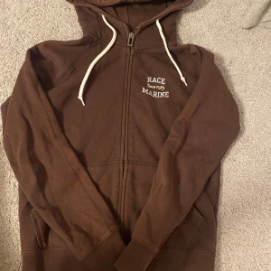 Brun hoodie från Race Marine - Säljer en brun hoodie från Race Marine i storlek 36. Den har en dragkedja framtill och en mysig huva med vita snören. Perfekt för kyliga dagar och har en klassisk stil som passar till det mesta. Använd men i bra skick!