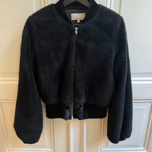 Svart bomberjacka i fuskpäls från Dry Lake X Vanessa Lindblad - Bra skick. Perfekt för höst och vinter med sin varma och fluffiga känsla. Jackan har en dragkedja framtill och långa ärmar. Den är både stilren och bekväm, idealisk för kyliga dagar.