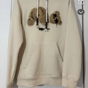 Beige hoodie från Palm Angels - Säljer en beige hoodie från Palm Angels med en unik broderad design på framsidan. Hoodien har en känguruficka och justerbara snören i huvan. Perfekt för en avslappnad stil.