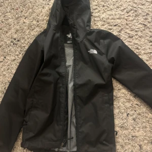 Svart regnjacka från The North Face - Säljer en svart regnjacka från The North Face med huva och dragkedja. Jackan har ett meshfoder för extra ventilation och komfort. Perfekt för regniga dagar och utomhusaktiviteter.