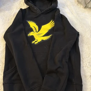 Svart hoodie från Lyle & Scott - Säljer en svart hoodie från Lyle & Scott med en stor gul örn på bröstet. Tröjan har en klassisk huva och en praktisk magficka. Perfekt för en avslappnad stil.🩷