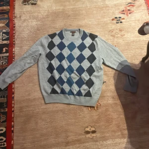 Grå tröja med diamantmönster från Dockers - Säljer en stilren grå tröja från Dockers med ett klassiskt diamantmönster i blått och mörkgrått. Tröjan har långa ärmar och rund halsringning, perfekt för en avslappnad men snygg look.