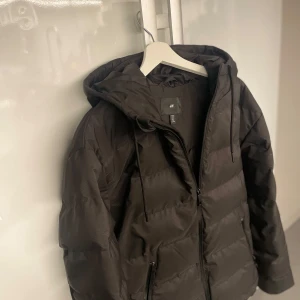 Svart pufferjacka med huva - Säljer en svart pufferjacka med huva. Jackan är vindtät och vattenavvisande, perfekt för kyliga dagar. Den har en dragkedja framtill och är långärmad. Passar bra för både höst och vinter. Nästan helt ny