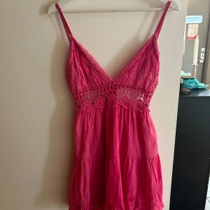 Rosa klänning med spetsdetaljer - Charmig rosa klänning med spetsdetaljer och smala axelband. Klänningen har en v-ringad fram och en öppen rygg med knytband. Perfekt för varma dagar.