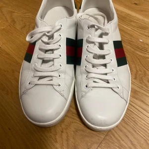 Gucci skor  - Snygga vita sneakers från Gucci med klassiska gröna och röda ränder på sidan. Skorna har snörning och en grön detalj med Gucci-logga på hälen. Perfekta för en stilren look. Säljer skorna eftersom dom har bara suttit i mitt rum. Köptes ganska länge sen utomlands. Dom är i använt skick men med inga defekter elr slitningar osv. Dom är äkta. Säljer dom väldigt billigt då jag inte har boxen elr kvitto på köpet. Men de som kan om skor kan lätt se att dessa är äkta. Pris kan diskuteras. Kp för frågor