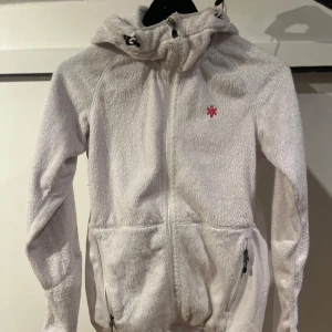 Vit fleecejacka med dragkedja - Mysig vit fleecejacka med dragkedja och huva från Áhkká. Jackan har två praktiska fickor med dragkedjor och en liten logotyp på bröstet. Perfekt för kyliga dagar. Storlek 34/36