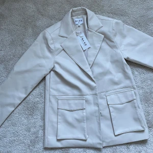 Beige kavaj från NA-KD 36 oversize oanvänd  - Snygg beige kavaj från NA-KD med klassisk krage och stora fickor framtill. Perfekt för en stilren look. Kavajen har en normal passform och är tillverkad i ett syntetmaterial.