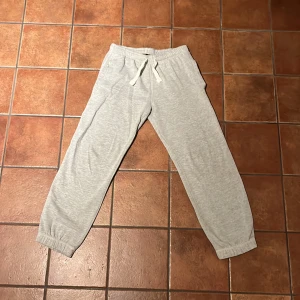 Grå mjukisbyxor från Lindex - Säljer ett par bekväma grå mjukisbyxor från Lindex. Byxorna har en elastisk midja med snörning och resår vid bensluten för extra komfort. Perfekta för en avslappnad dag hemma.