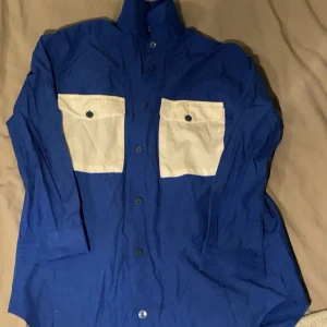 Blå skjorta med beige fickor - Snygg blå skjorta med kontrasterande beige fickor framtill. Skjortan har långa ärmar och knappar hela vägen ner. Perfekt för en stilren look.